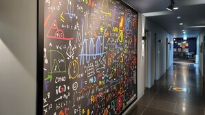 Un couloir où est accroché un grand tableau noir, couvert d’équations et de schémas mathématiques et scientifiques aux couleurs vives.