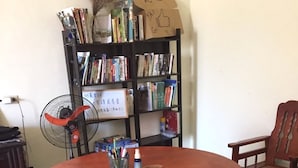 Une table ronde en bois, entourée de quatre tabourets, avec une bibliothèque remplie de livres et un ventilateur à proximité.
