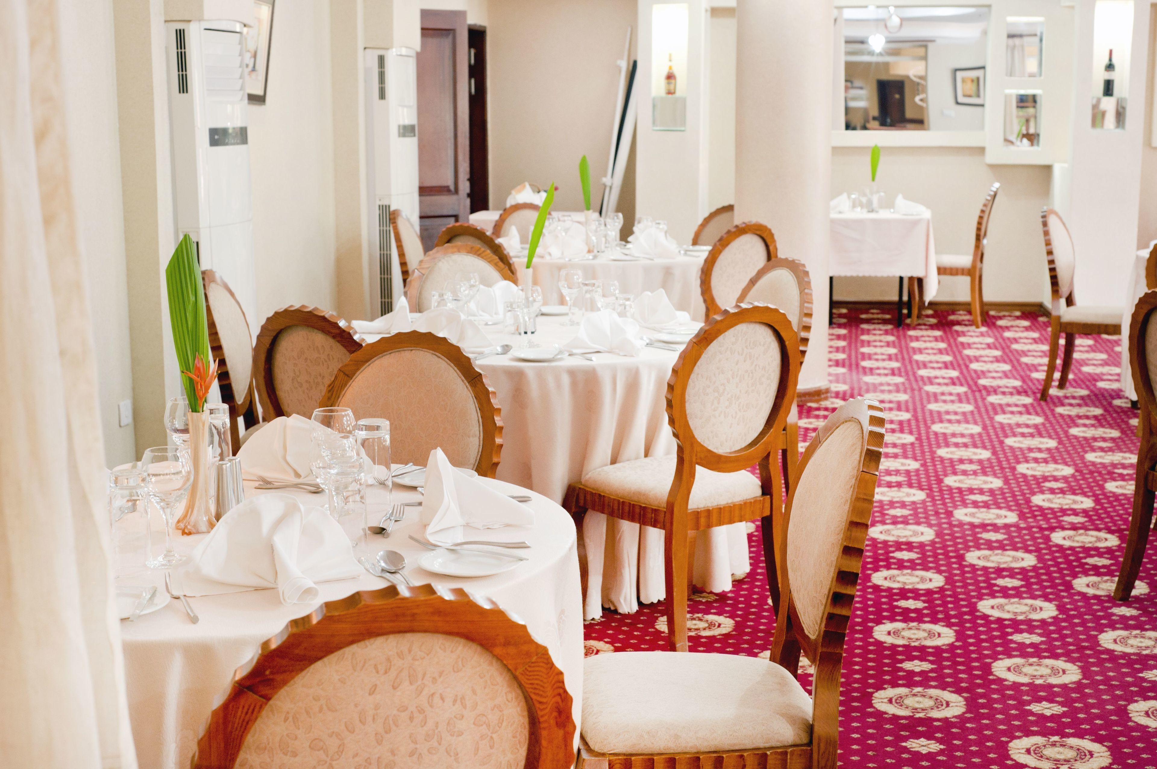 A dining area with round tables set for multiple guests, each with white tablecloths and neatly arranged cutlery.