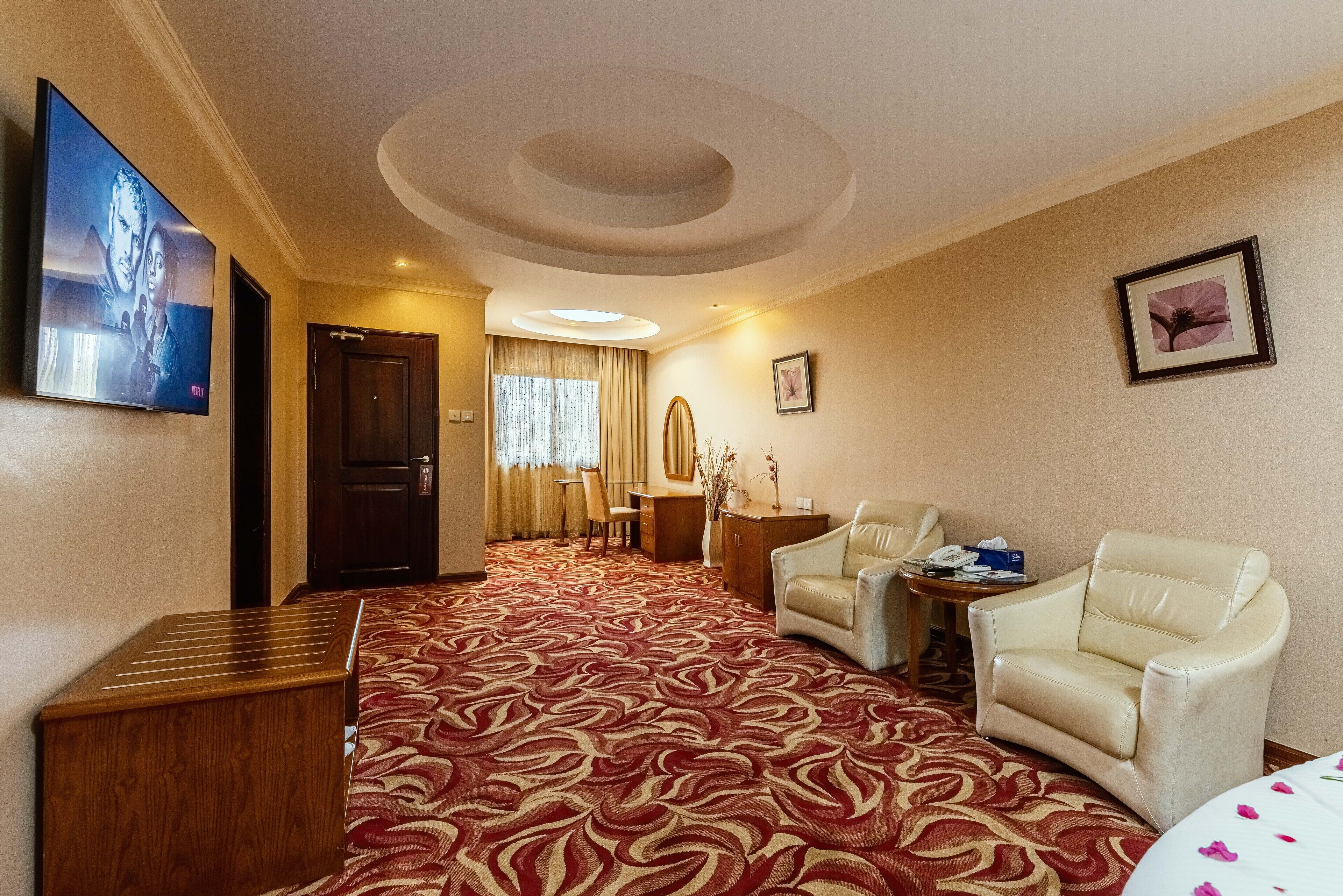 A hotel room with a red patterned carpet, a television mounted on the wall, a desk with a chair, a sofa, and a small table with a vase.