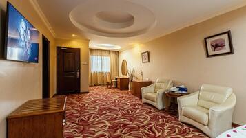 A hotel room with a red patterned carpet, a television mounted on the wall, a desk with a chair, a sofa, and a small table with a vase.