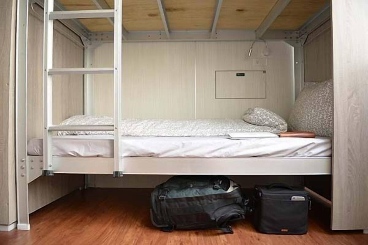 Una cama litera con colchón, almohadas y una mesita de noche pequeña. Se ven una maleta y un bolso en el suelo.