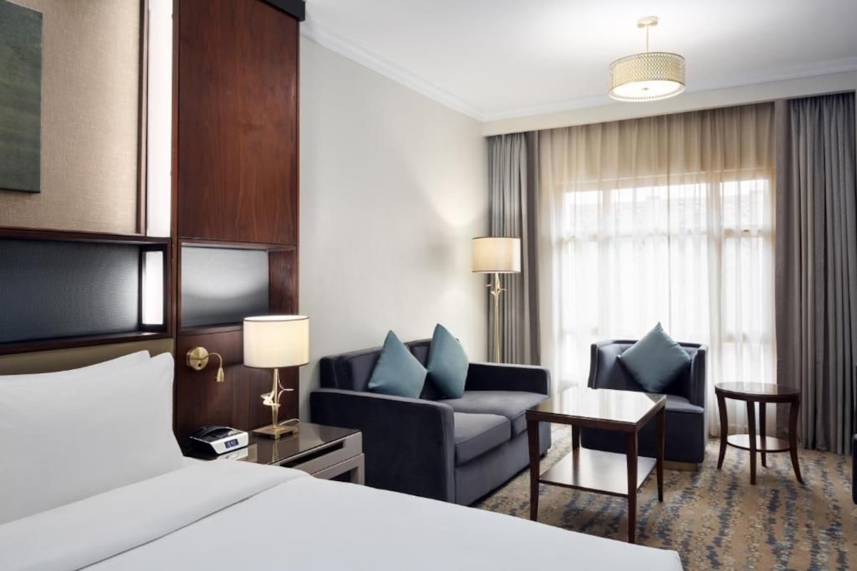 A hotel room with a bed, two sofas, a small table, a lamp, and a large window with curtains.