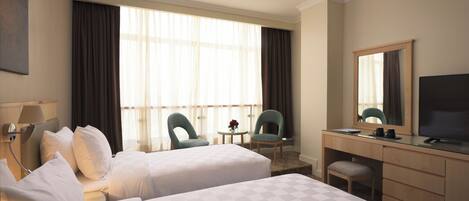 A hotel room with two beds, a desk, a chair, a TV, and a large window with curtains.
