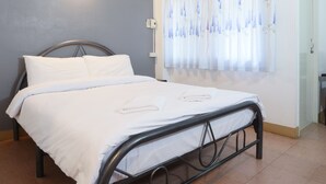 A neatly made bed with white linens in a room with a grey wall, a window with floral curtains, and a door to the right. - Pakdee House (Udon Thani)