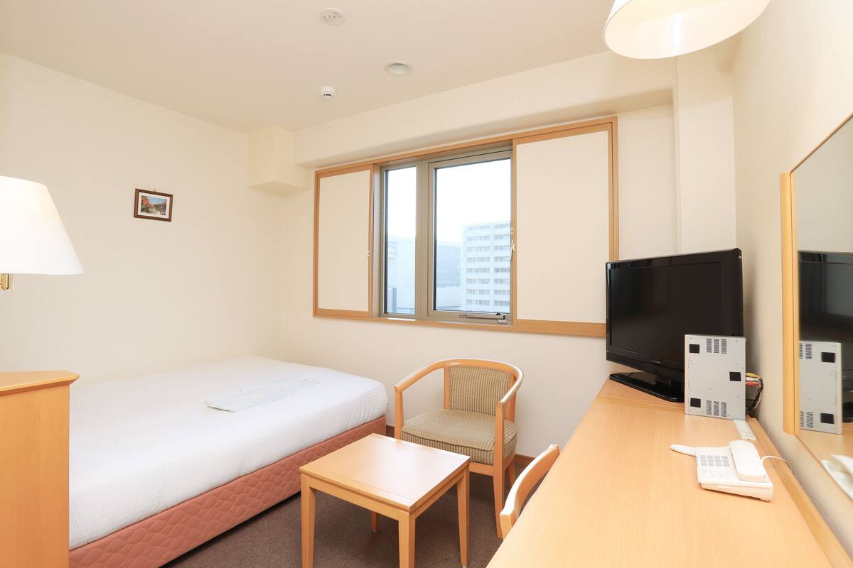 A hotel room with a single bed, a desk with a chair, a television, and a window with a view of buildings.