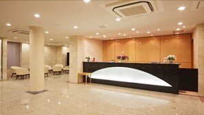 Zona de recepción de un hotel con un mostrador curvo, varias sillas y suelo de baldosas.