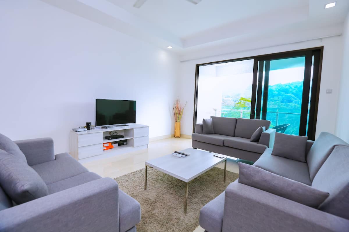 A modern living room with a flat-screen TV, grey sofas, a coffee table, and a large sliding glass door leading to a balcony with a view of greenery.