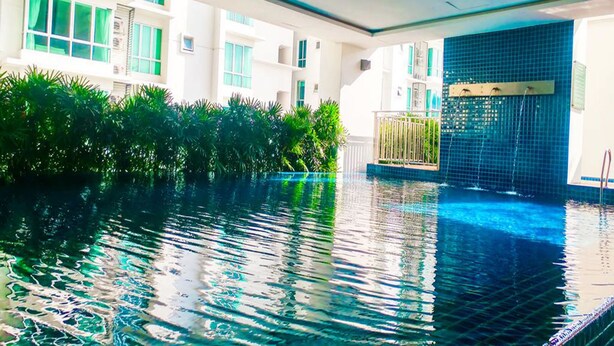 Une piscine dont le bord est revêtu de carreaux, entourée d’une végétation luxuriante et bordée d’un bâtiment doté de nombreuses fenêtres.