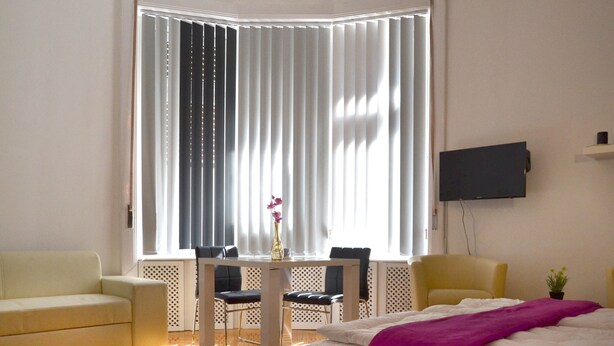 Una habitación de hotel moderna con un ventanal grande, una cama, un sofá, una mesa de comedor con sillas y un televisor montado en la pared.