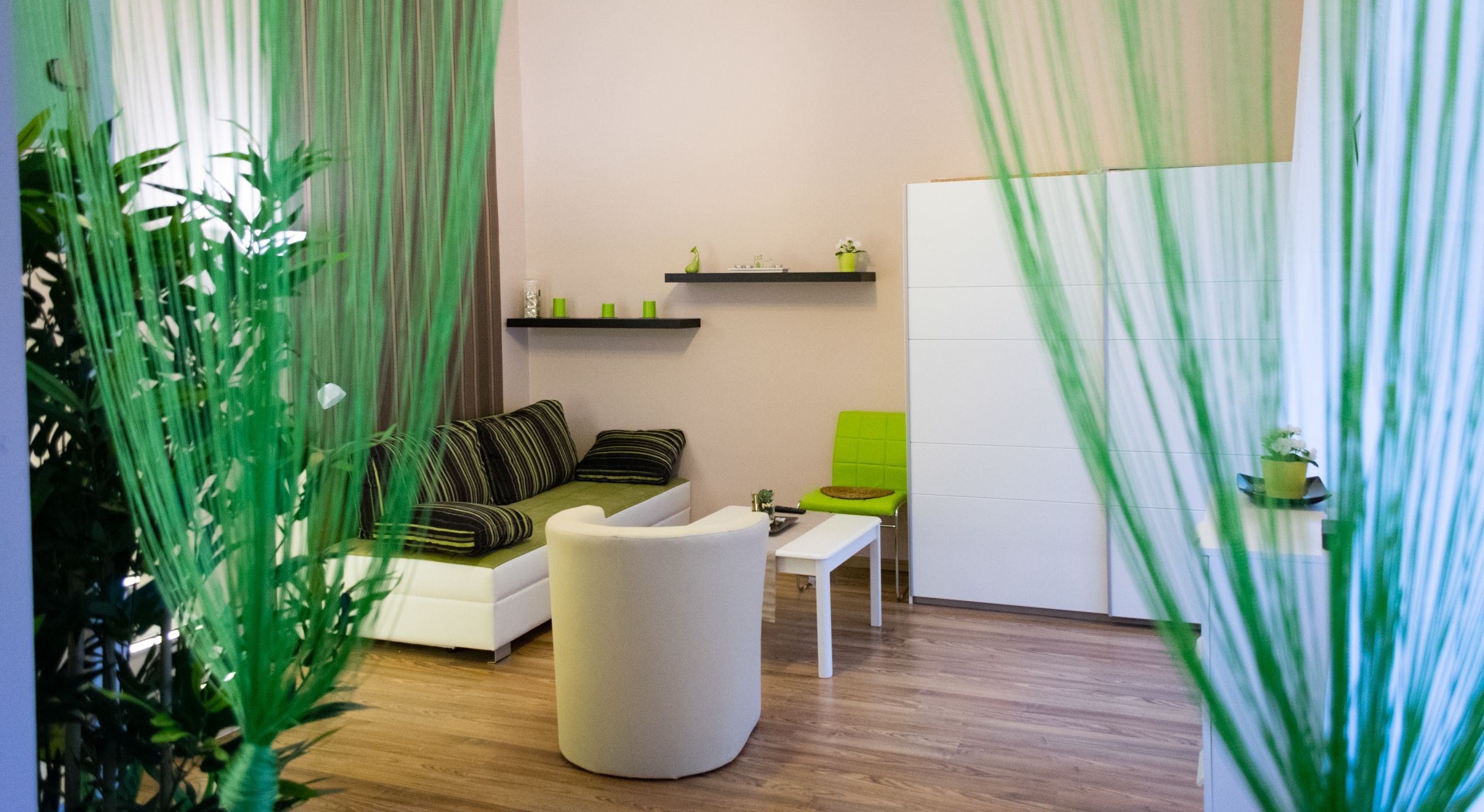 A modern living room with a sofa, a white armchair, a small table, and a green chair. There are shelves with decorative items and a potted plant on the wall.