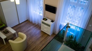 Una habitación de hotel moderna con piso de madera, una mesita de noche blanca, una silla amarilla, un tocador blanco, un televisor de pantalla plana montado en la pared y un balcón de vidrio con vista a la ciudad.
