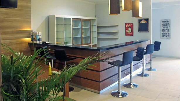 A modern reception area with a wooden counter, bar stools, and a large plant. - The Alley City Hotel (Denpasar)