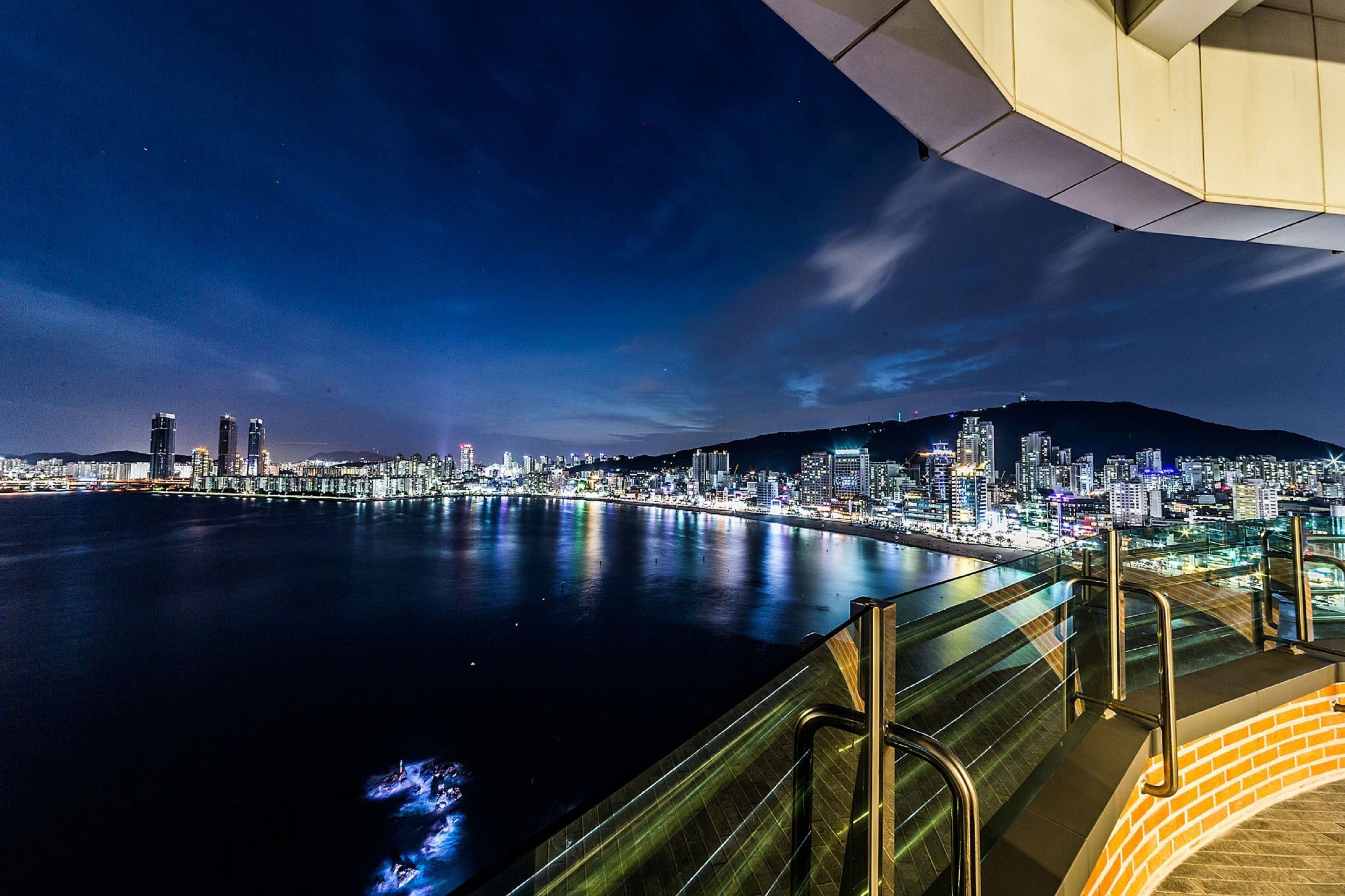 A nighttime cityscape with a waterfront view, featuring illuminated buildings and a bridge.