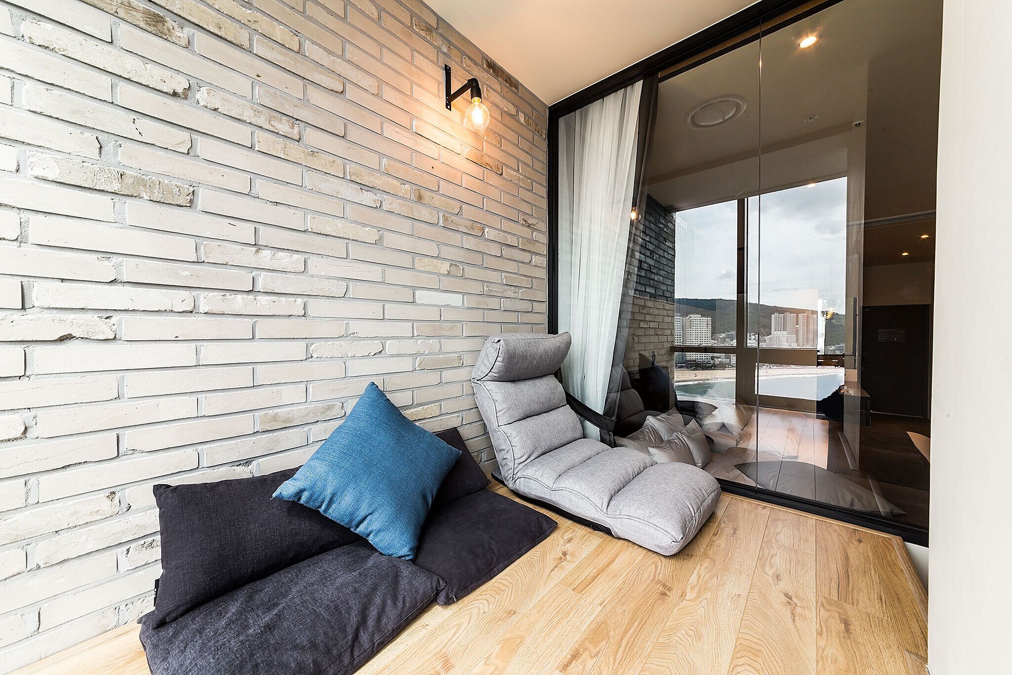 A modern room with a brick wall, a sofa with cushions, a recliner, and a large window offering a view of the city.