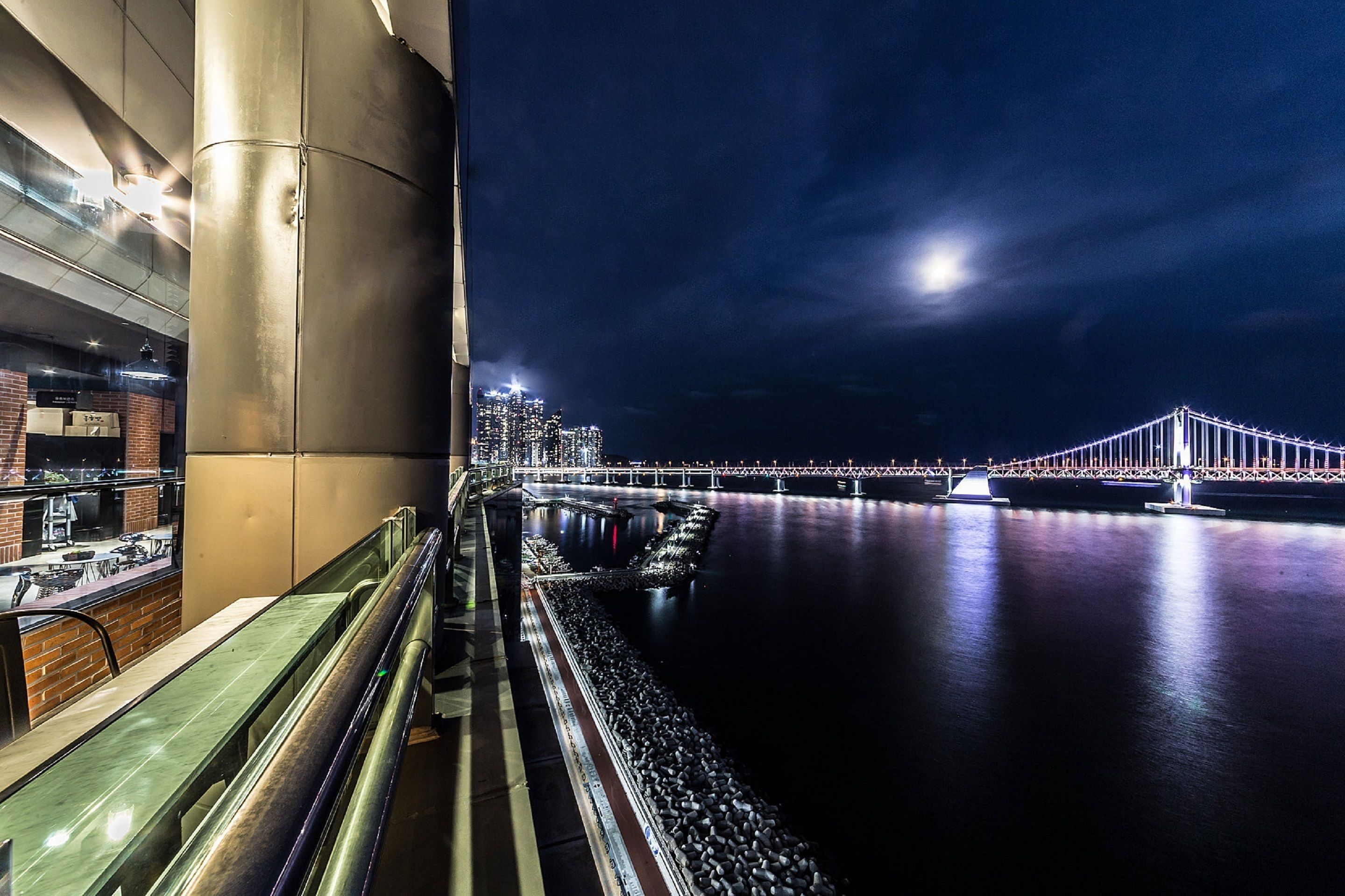A nighttime view of a river with a bridge, a building with a glass facade, and a railing along the riverbank.