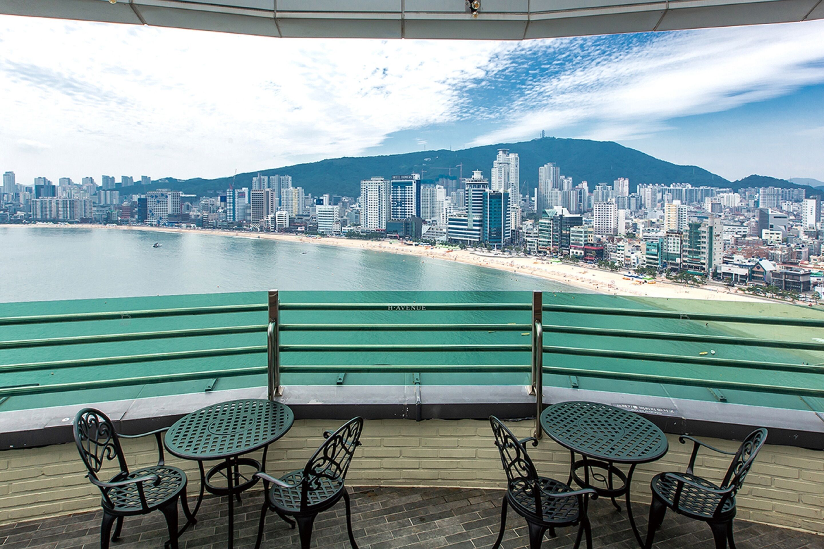 A terrace with green railings and tables overlooking a cityscape with a beach and mountains.