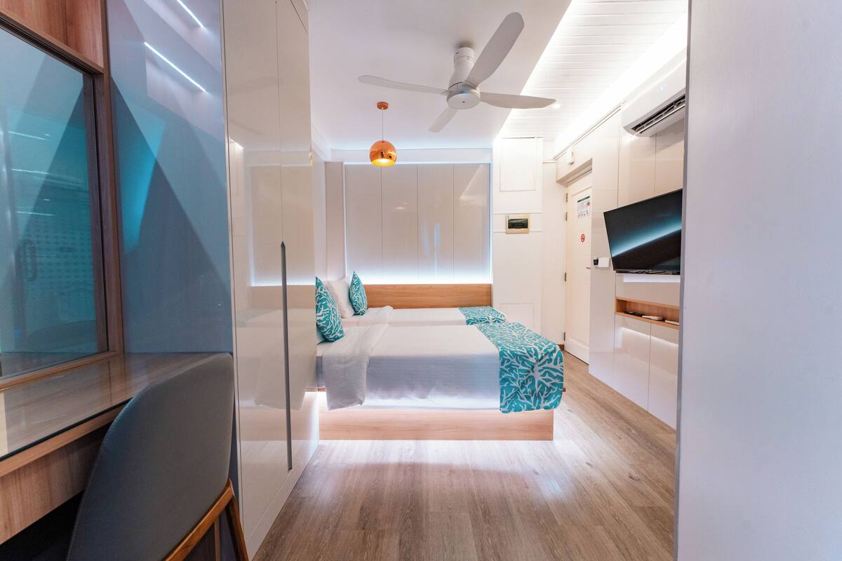 A modern bedroom with a bed, a desk, a chair, a ceiling fan, and a TV.