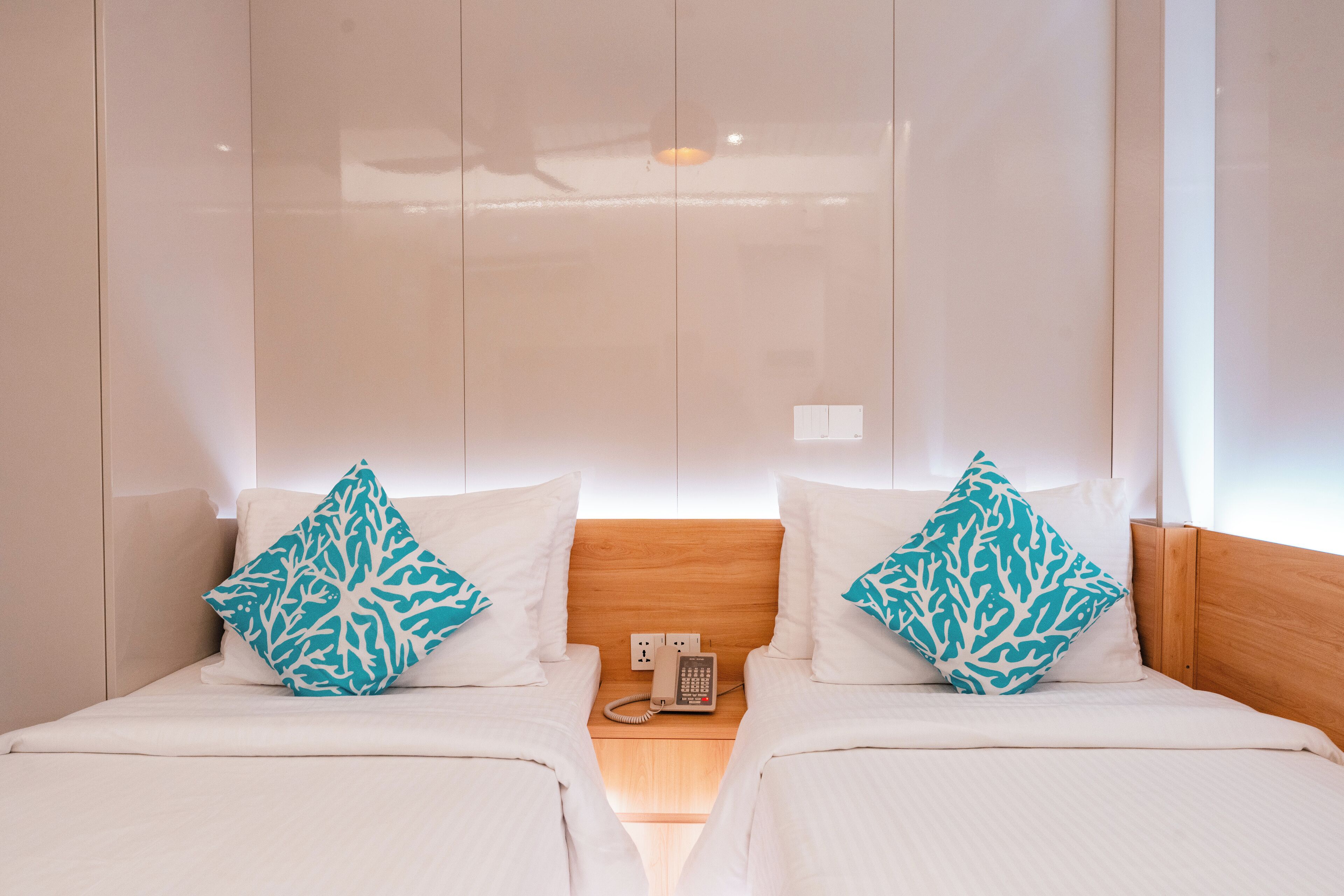 Habitación de hotel con dos camas, cada una con ropa de cama blanca y almohadas teal estampadas. Tiene un cabecero de madera, una mesita de noche con teléfono y una lámpara de pared.