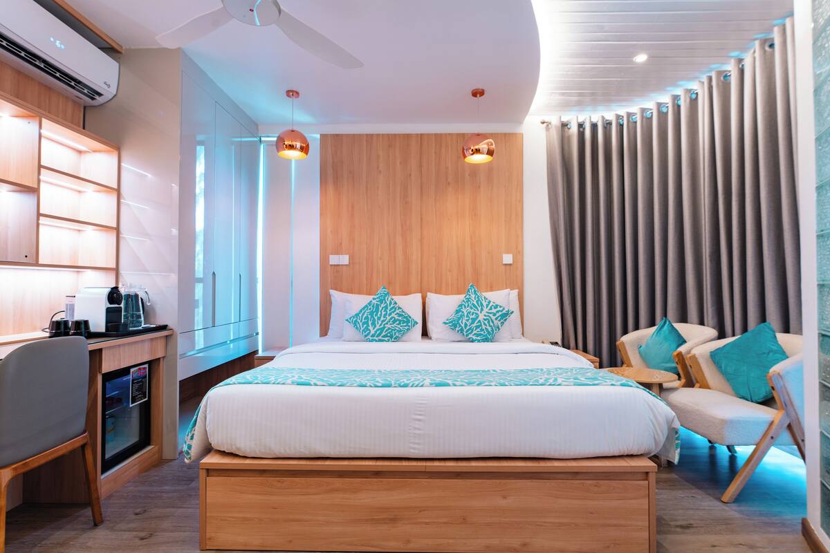 A modern hotel room with a wooden headboard, a large bed with patterned bedding, a desk with a coffee maker, and a seating area with chairs.