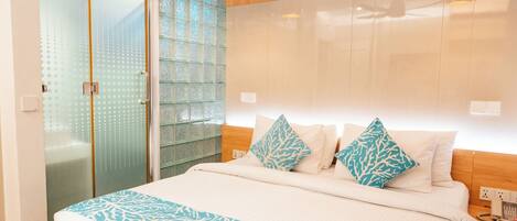 Quarto de hotel moderno com uma cama grande, box de vidro e cabeceira de madeira.