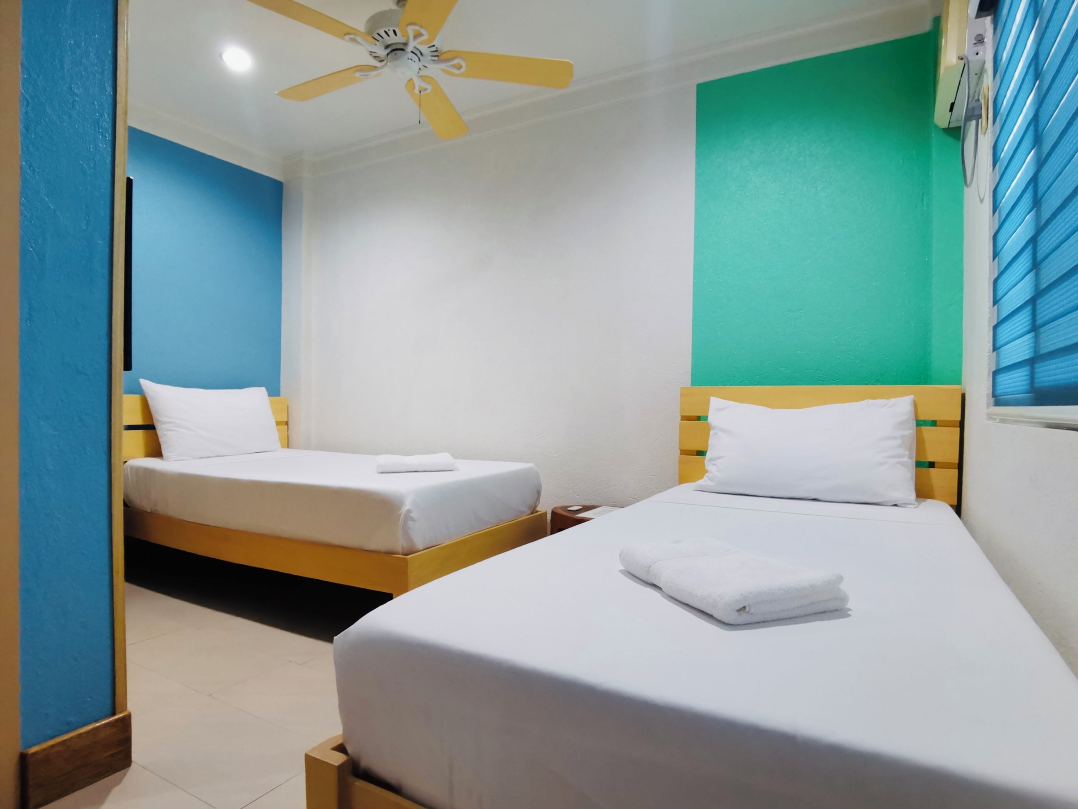 Habitación de hotel con dos camas, ventilador de techo y ventana con persianas.