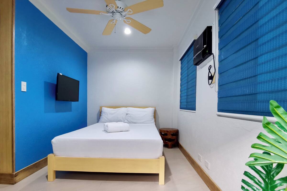 A bedroom with a bed, a ceiling fan, a mounted TV, and a window with blinds.