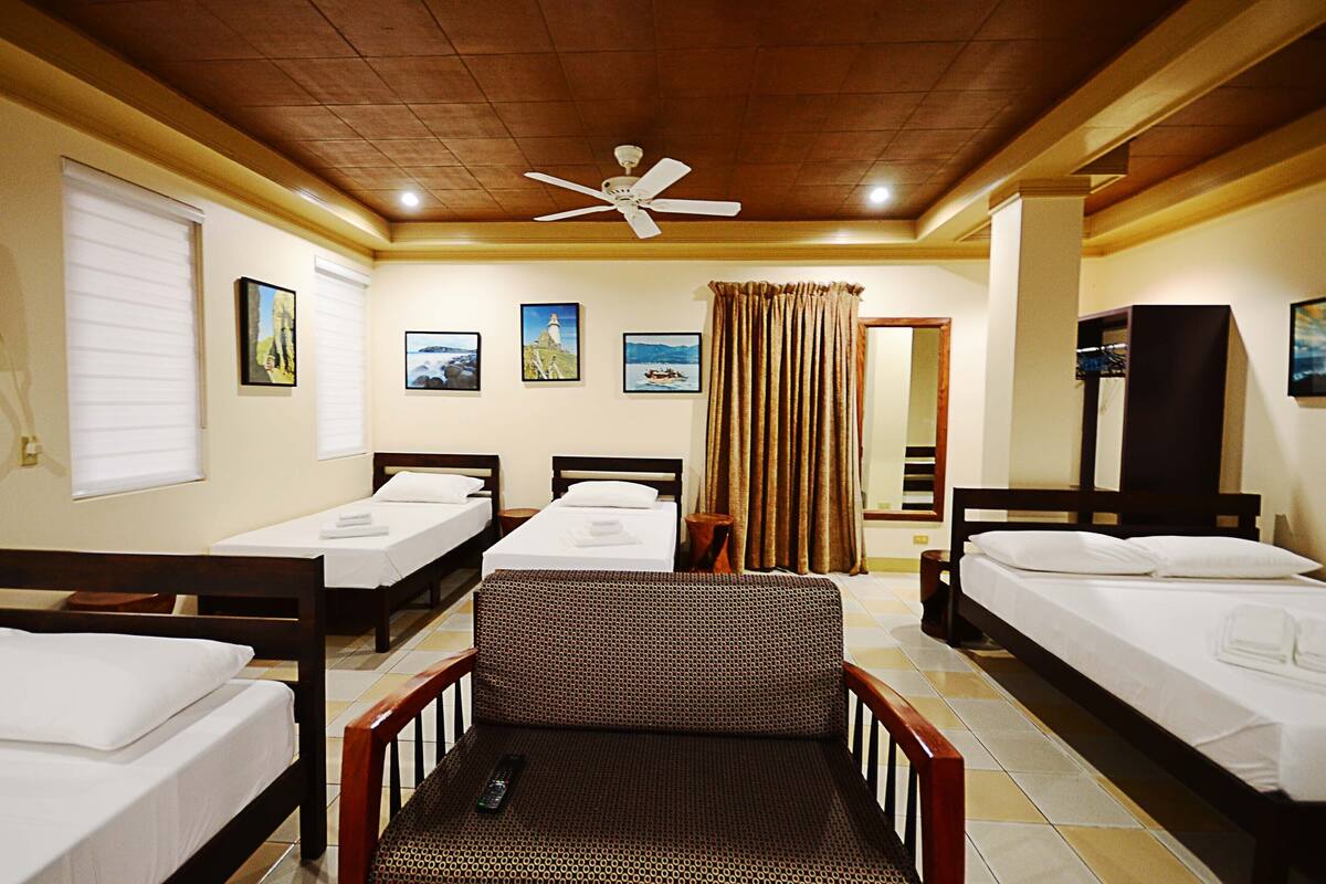 A hotel room with three single beds, a sofa, a ceiling fan, and a window with blinds.