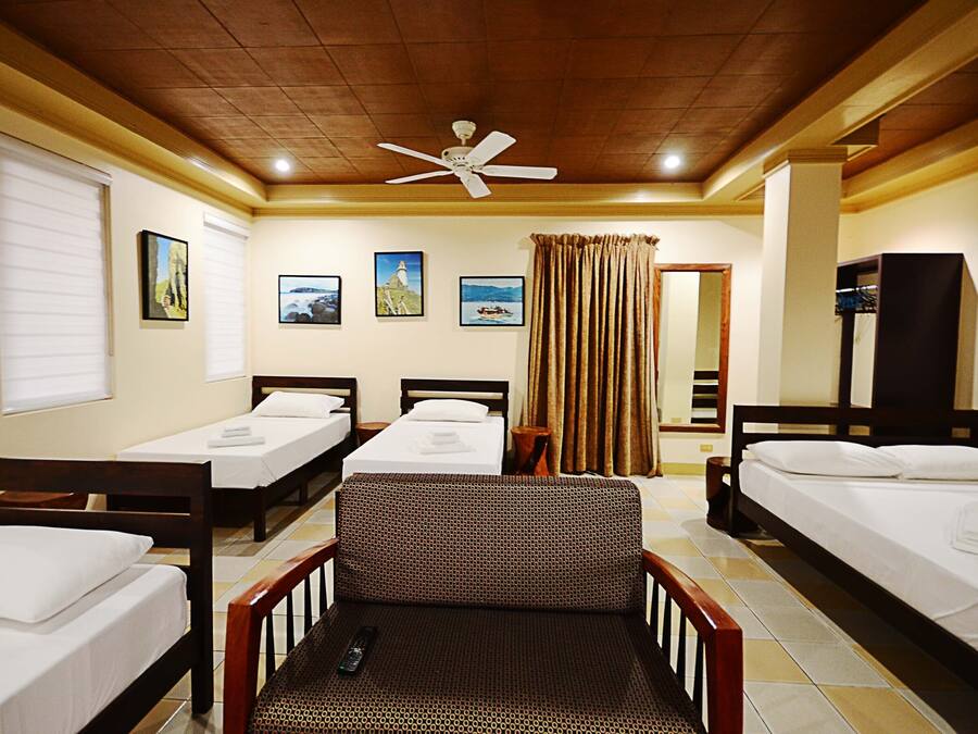 A hotel room with three single beds, a sofa, a ceiling fan, and a window with blinds.