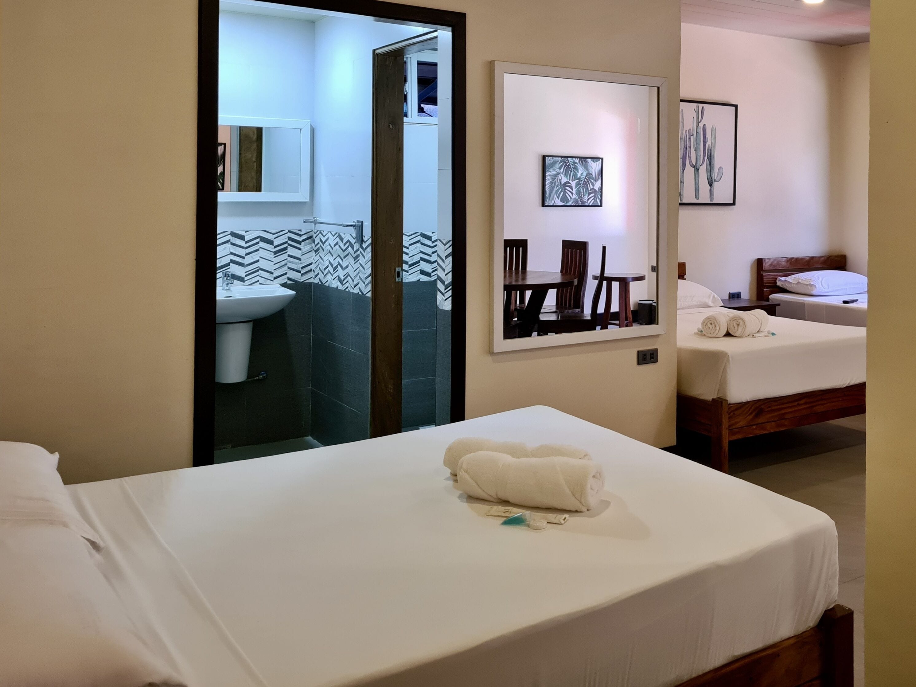 Habitación de hotel con cama, baño con lavamanos y espejo, y zona de comedor con sillas y mesa.