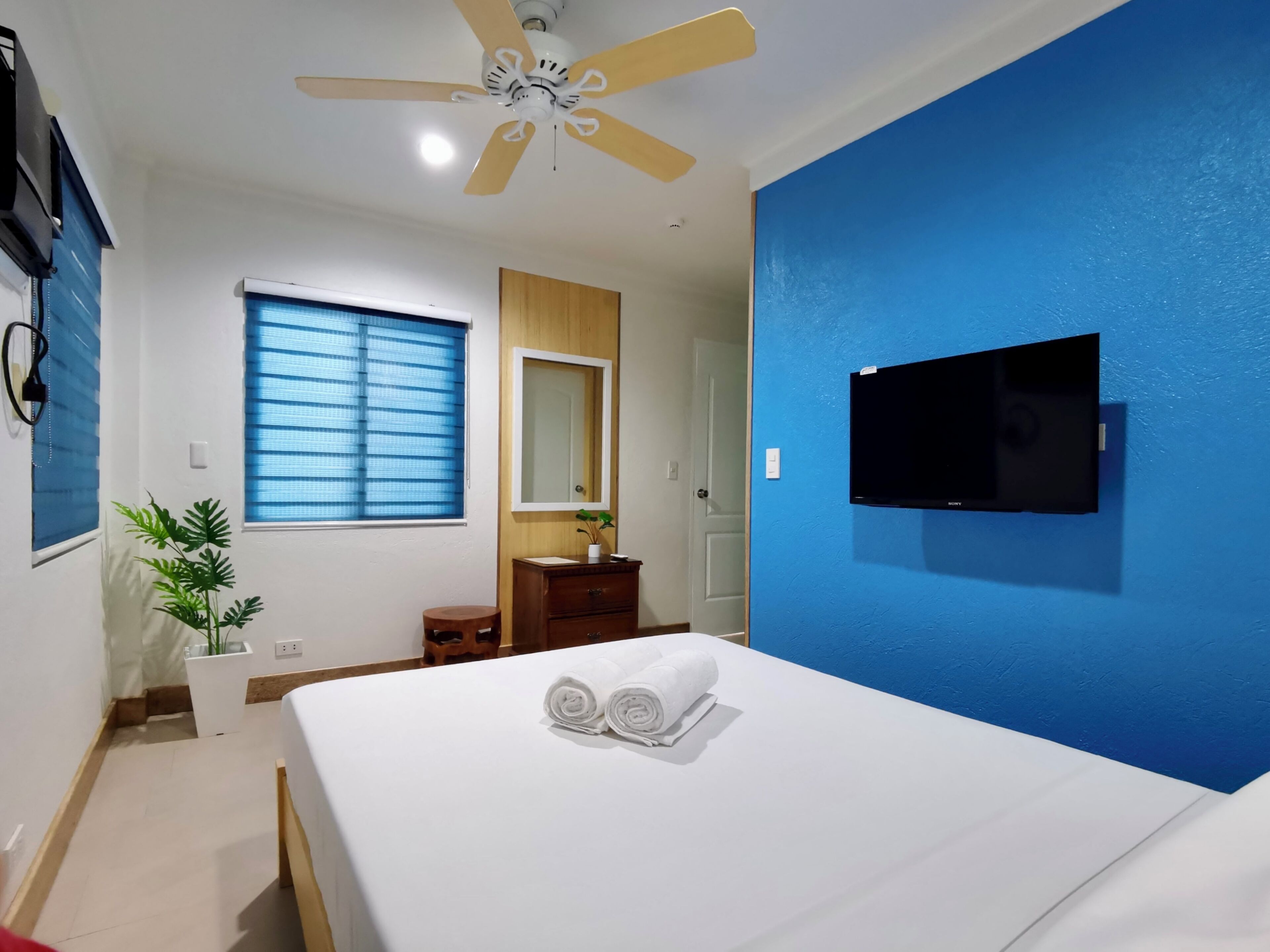 Habitación de hotel con cama, ventilador de techo, televisor montado en la pared, ventana con persianas, aparador de madera y planta en maceta.