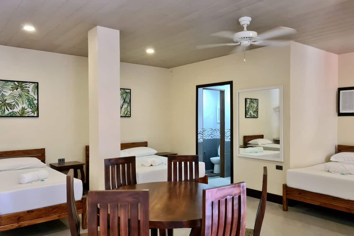 A hotel room with two beds, a dining table, and chairs. There is a ceiling fan and a large mirror on the wall.