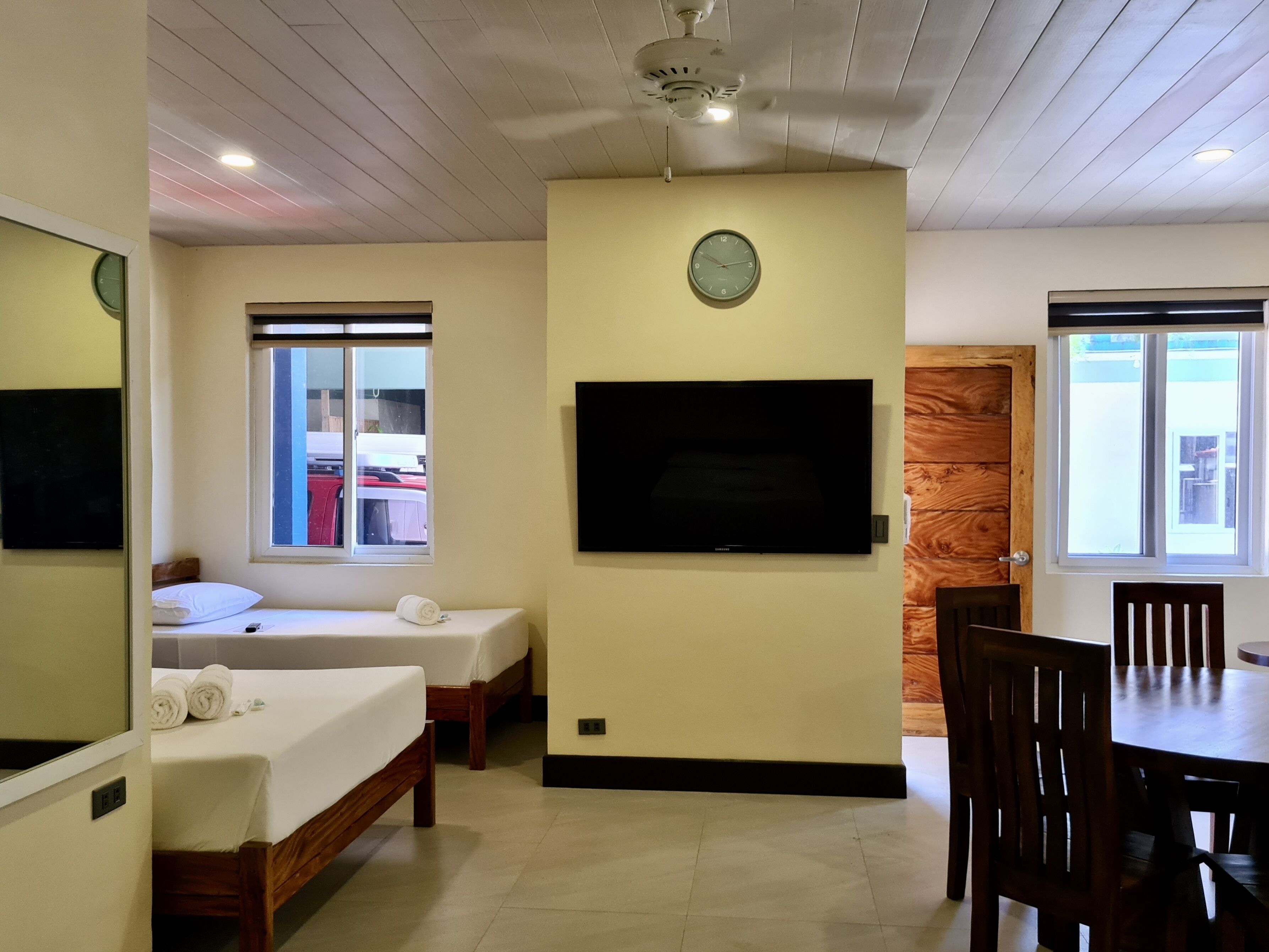 A hotel room with two beds, a television, a clock, and a dining area with wooden chairs.