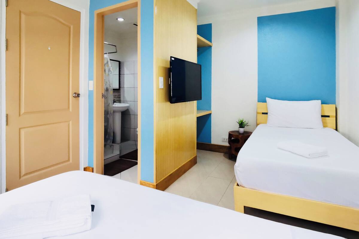 A hotel room with two beds, a TV, a small table with a plant, and a bathroom visible through an open door.