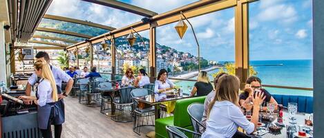 A rooftop restaurant with outdoor seating, overlooking a coastal cityscape.
