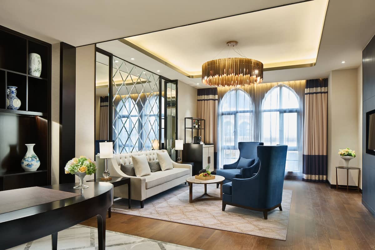 Una sala de estar moderna con un sofá, un sillón y una mesa de centro, que cuenta con una araña, ventanas amplias y una estantería.