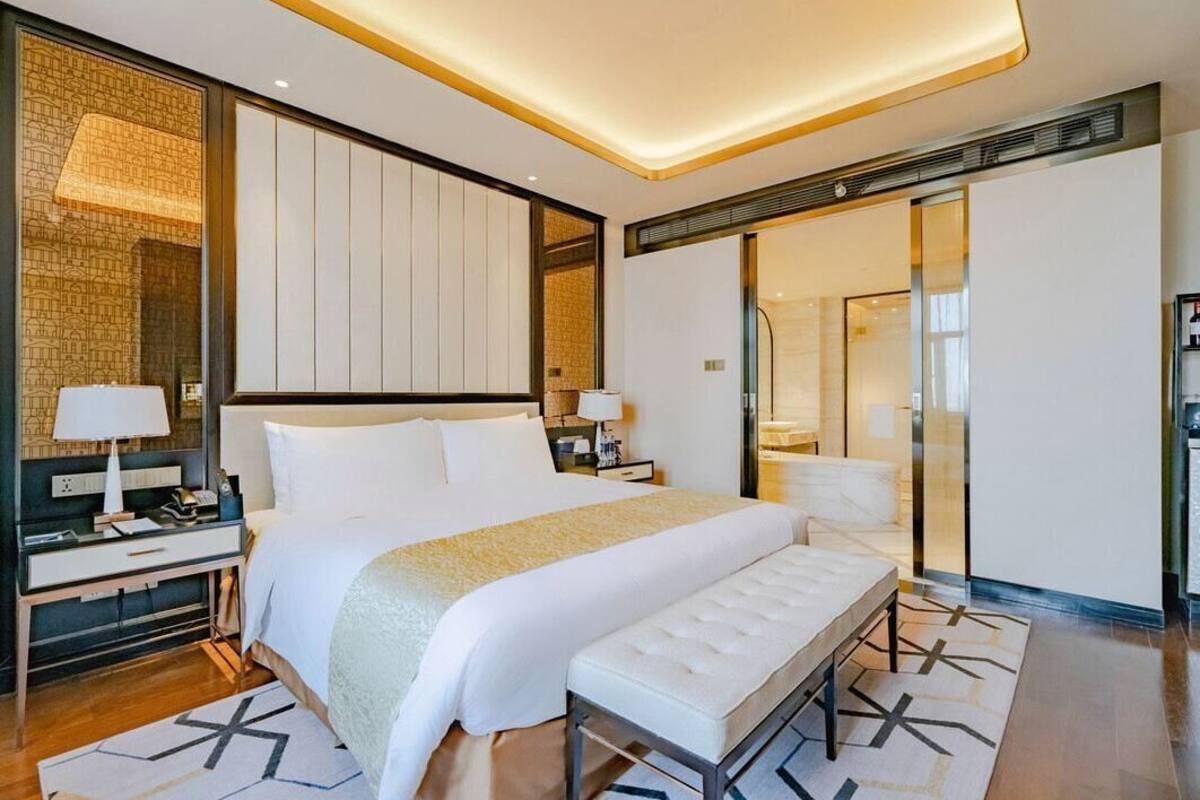 Una habitación de hotel moderna con una cama grande, mesitas de noche, un banco y un espejo amplio.