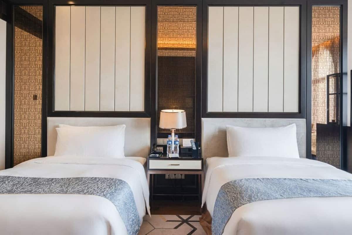 Habitación de hotel con dos camas, una mesita de noche, una lámpara y un papel tapiz estampado.