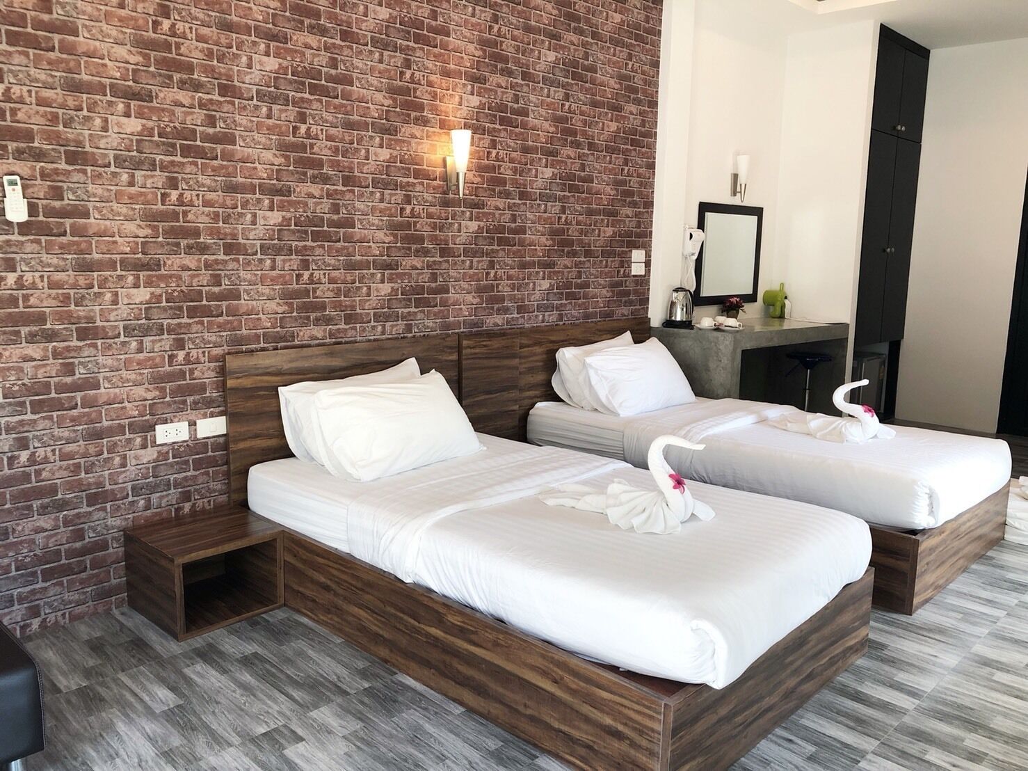A hotel room with two wooden beds, a brick wall, and a small desk with a mirror and a kettle.