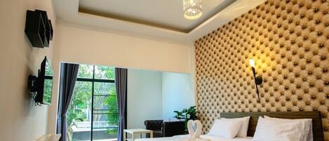 A hotel room with a large bed, a tufted headboard, a television mounted on the wall, and a view of greenery through the window.