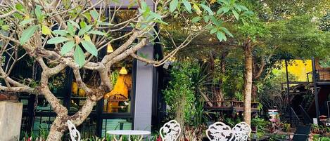 An outdoor seating area with white metal chairs and tables, surrounded by lush greenery and a tree.