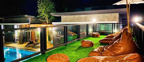 A modern outdoor pool area with artificial grass, leather bean bags, and a glass-enclosed building.