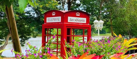 Una cabina telefónica roja con texto en tailandés, rodeada de plantas y árboles coloridos.