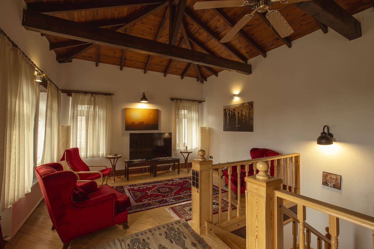 Un salón amplio con techo de madera alto, un televisor de pantalla plana grande, sillones tapizados en rojo, una escalera de madera y una alfombra estampada.