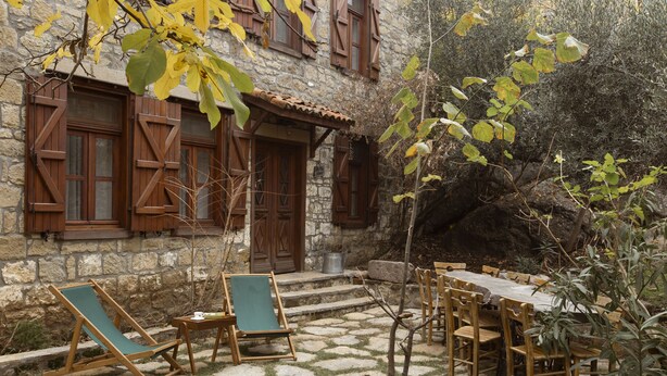 A stone building with wooden shutters, a stone patio with a table and chairs, and a small garden area. - Mavras Tas Odalar (Ayvacik)