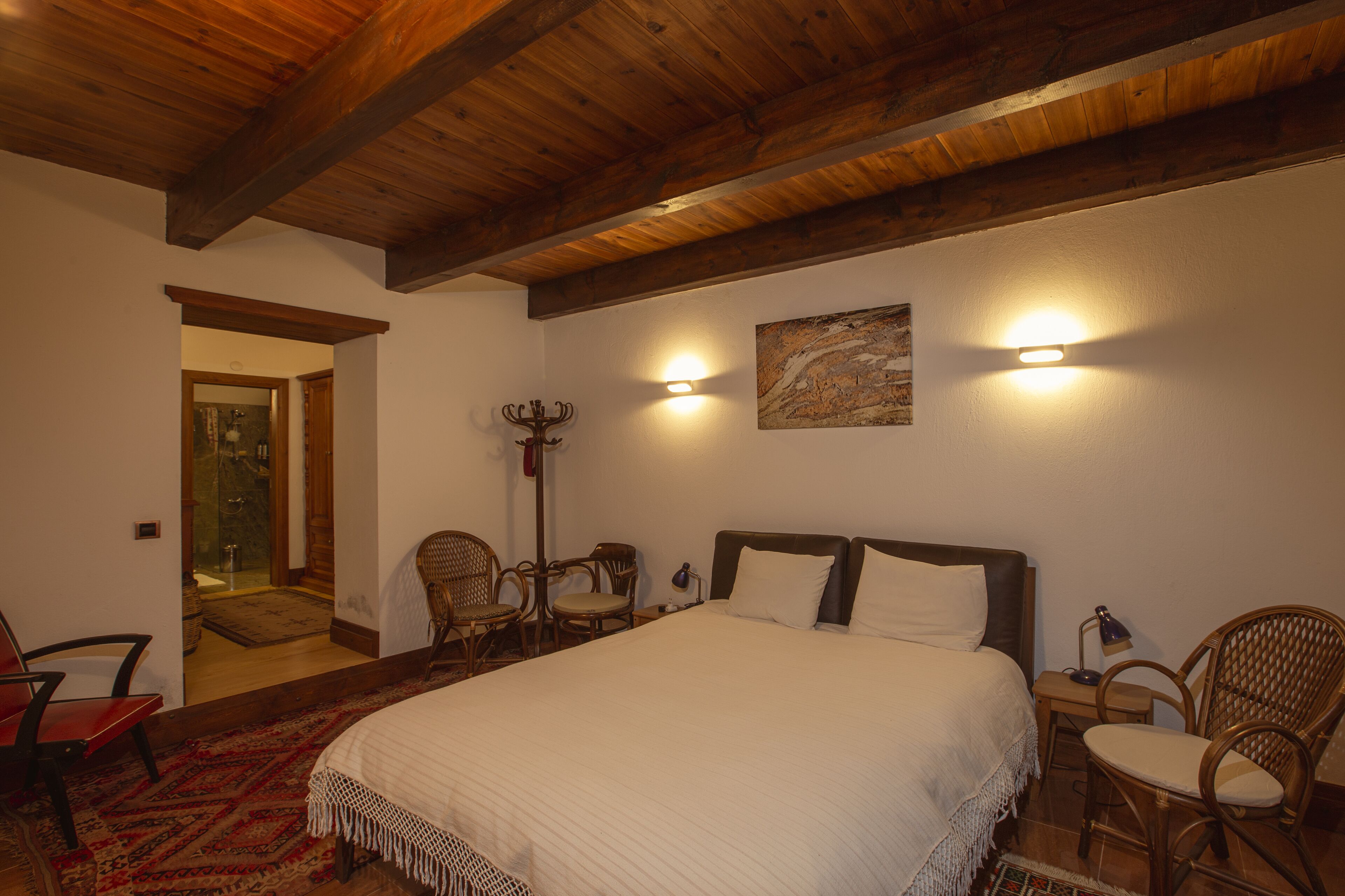 Un dormitorio con una cama grande, vigas de madera en el techo, dos luces fijadas a la pared, un cuadro y sillas de mimbre.