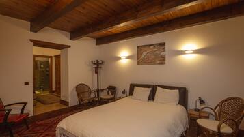 Un dormitorio con una cama grande, vigas de madera en el techo, dos luces fijadas a la pared, un cuadro y sillas de mimbre.