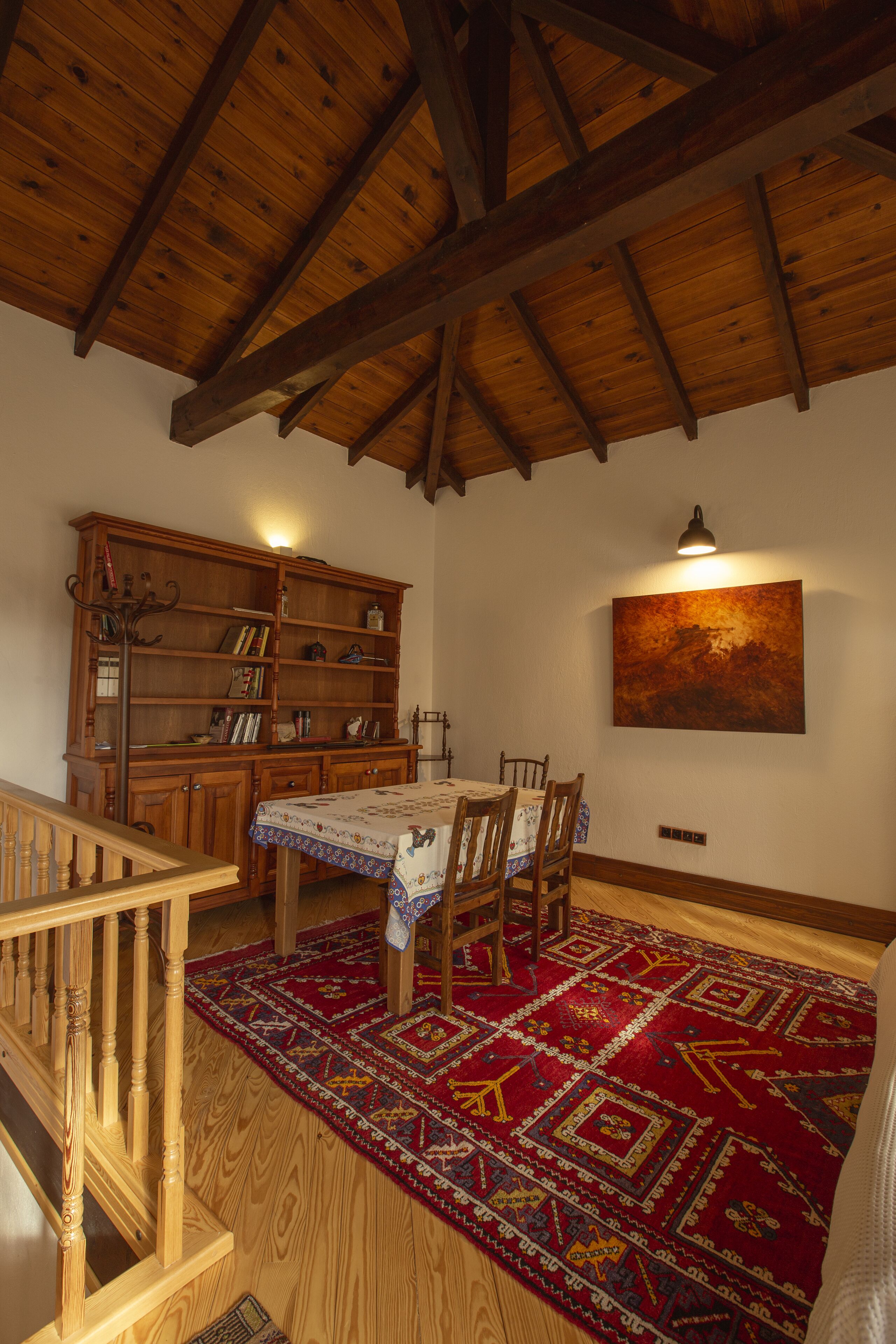 A dining area with a wooden ceiling, a large bookshelf, a table with chairs, and a patterned rug.