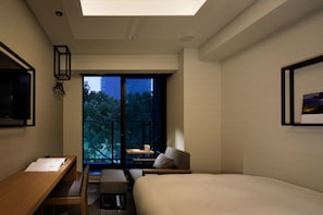 Habitación de hotel moderna con una cama grande, un escritorio, una silla y un balcón con vistas a árboles y edificios.
