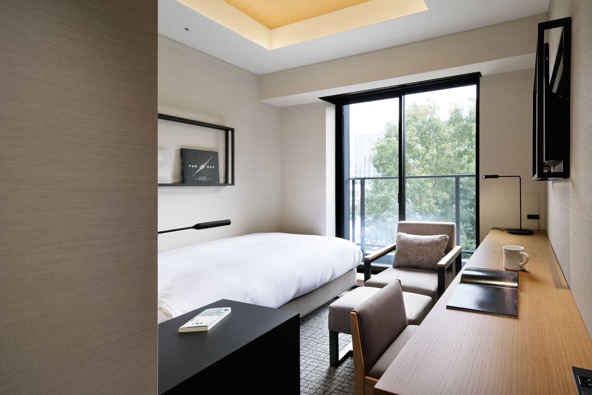 Habitación de hotel moderna con una cama, un escritorio, una silla, una taza de café y un amplio ventanal con vistas a árboles.